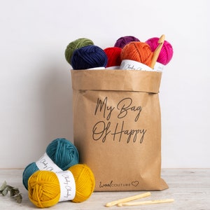 May include: A brown paper bag filled with colorful yarn balls and crochet hooks. The bag has the text "My Bag Of Happy" printed on it. The yarn balls are in various colors, including red, orange, yellow, green, blue, and purple. The crochet hooks are made of wood and are in a natural color. The bag is sitting on a wooden surface.