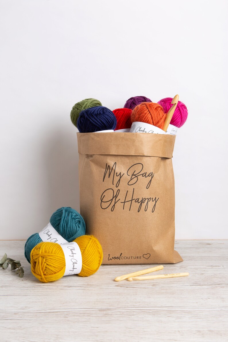 May include: A brown paper bag filled with colorful yarn balls and crochet hooks. The bag has the text "My Bag Of Happy" printed on it. The yarn balls are in various colors, including red, orange, yellow, green, blue, and purple. The crochet hooks are made of wood and are in a natural color. The bag is sitting on a wooden surface.
