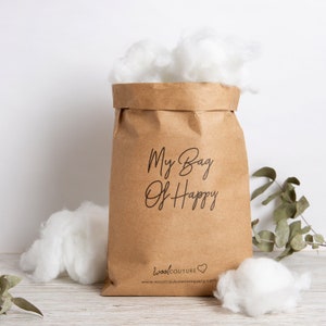 May include: A brown paper bag filled with fluffy white material, with the words "My Bag Of Happy" printed on the front. The bag is surrounded by more of the white material, and the Wool Couture Company logo is at the bottom.