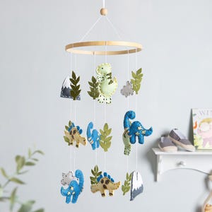 May include: A hanging nursery mobile featuring felt dinosaur figures in shades of blue, green, and cream, interspersed with felt leaves, clouds, and mountain shapes. The figures are suspended from a circular wooden frame by thin white strings.