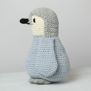 May include: A crocheted penguin toy with a gray head, white face, and light blue body. The penguin is standing on a white surface.