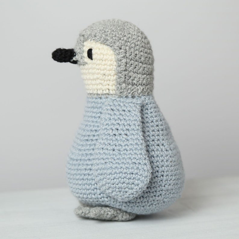 May include: A crocheted penguin toy with a gray head, white face, and light blue body. The penguin is standing on a white surface.