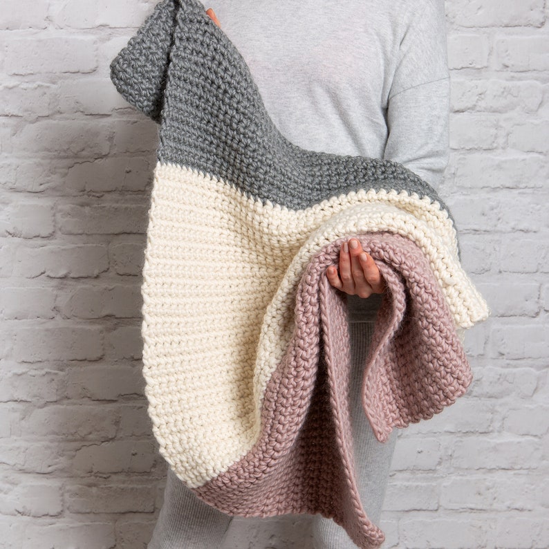 May include: A crocheted throw blanket in shades of gray, cream, and pink. The blanket is folded in half and held by a person.