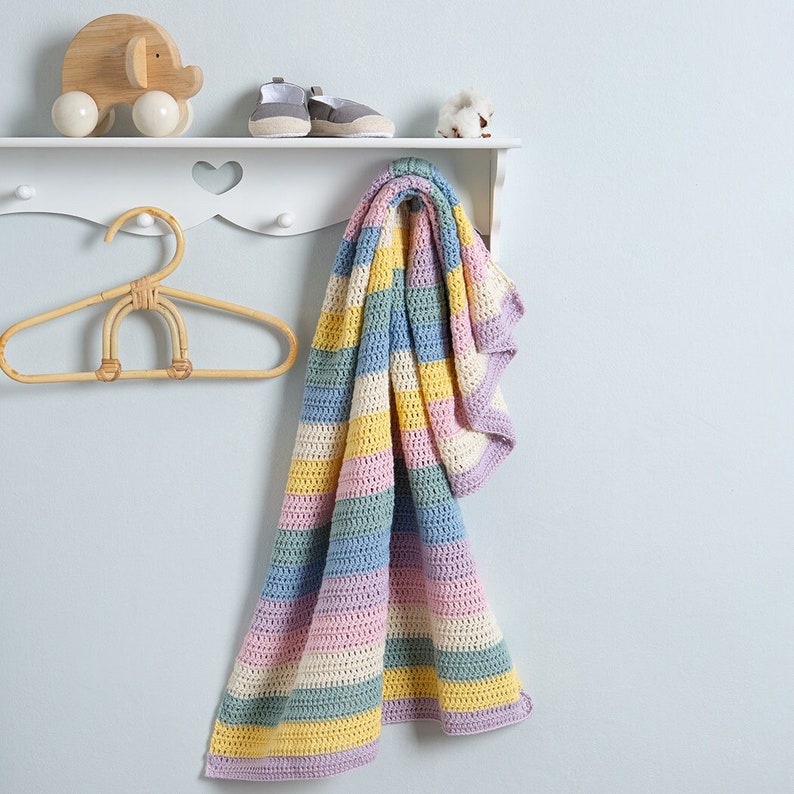 May include: A crocheted blanket featuring horizontal stripes in pastel yellow, light blue, soft pink, and sage green. The blanket is draped over a white wooden wall shelf that also holds a wooden elephant toy, gray baby shoes, and a cotton boll.