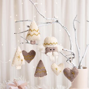 May include: A collection of seven knitted hanging ornaments in cream and taupe colors, featuring shapes of Christmas trees, hearts, and a bearded figure. Each piece is detailed with gold embroidered accents and hangs from a thin white string.