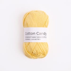 May include: A skein of pale yellow yarn with a white paper label reading Cotton Candy, DK weight yarn, 100% cotton, 50 grams, 125 meters. The yarn is wound into an oval shape with visible strands of soft, light yellow fiber throughout the entire ball.