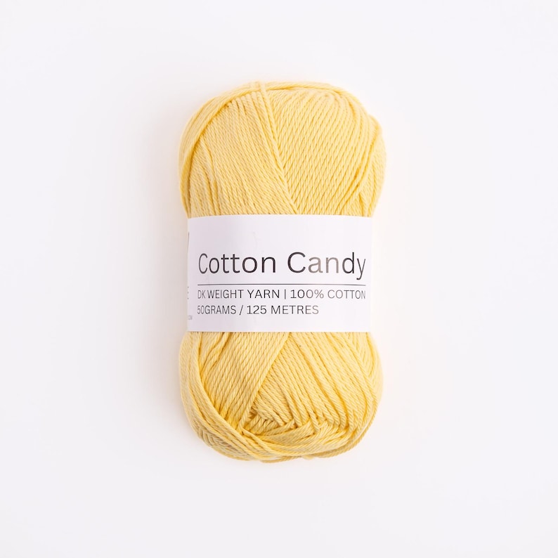 May include: A skein of pale yellow yarn with a white paper label reading Cotton Candy, DK weight yarn, 100% cotton, 50 grams, 125 meters. The yarn is wound into an oval shape with visible strands of soft, light yellow fiber throughout the entire ball.