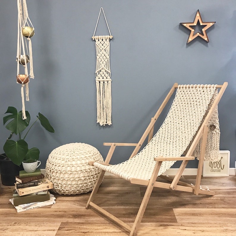 May include: A white macrame chair with a wooden frame, a white macrame wall hanging, a white knitted pouf, and a star-shaped light fixture on a blue wall. The wall also has a sign that says "Wood Couture".