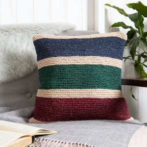 K&ouml;nnte beinhalten: Ein quadratisches Strickkissen mit horizontalen Streifen in Marineblau, Creme, Waldgr&uuml;n und Dunkelburgunder. Die strukturierte Garnverarbeitung erzeugt ein gleichm&auml;&szlig;iges kraus rechtes Strickmuster &uuml;ber die gesamte Oberfl&auml;che des Dekokissens.