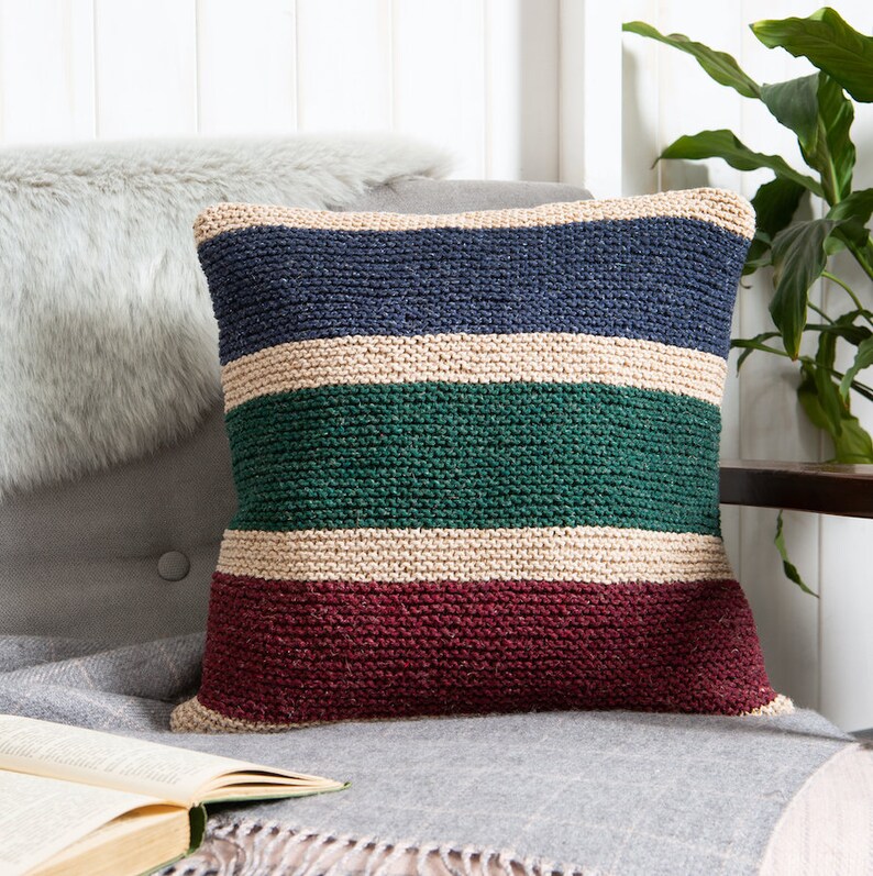 K&ouml;nnte beinhalten: Ein quadratisches Strickkissen mit horizontalen Streifen in Marineblau, Creme, Waldgr&uuml;n und Dunkelburgunder. Die strukturierte Garnverarbeitung erzeugt ein gleichm&auml;&szlig;iges kraus rechtes Strickmuster &uuml;ber die gesamte Oberfl&auml;che des Dekokissens.
