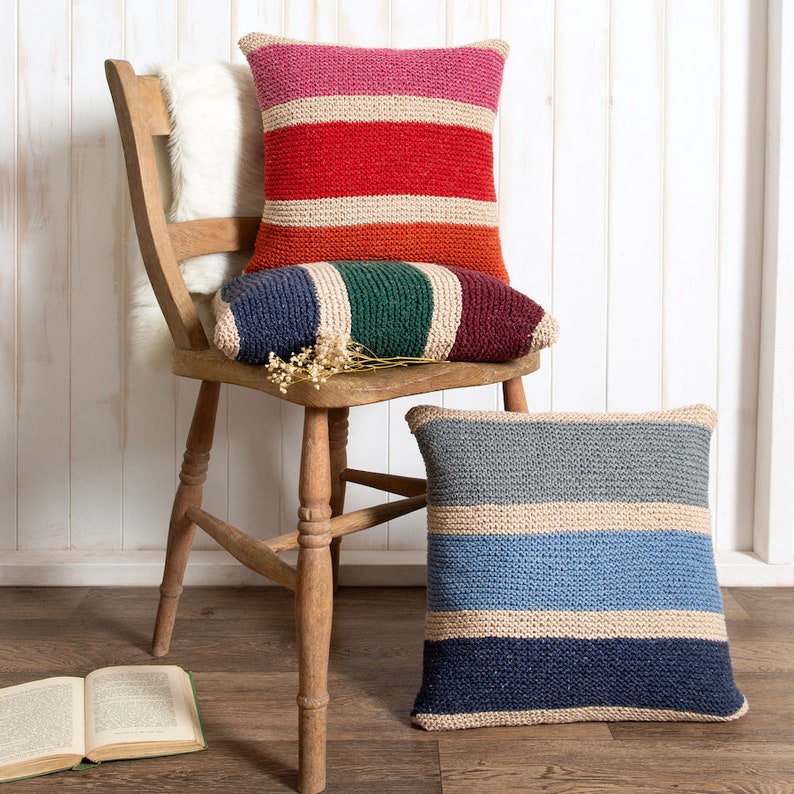 K&ouml;nnte beinhalten: Drei quadratische Strickkissen mit horizontalen Streifen. Ein Kissen zeigt rosa, beige, rote und orangefarbene B&auml;nder. Ein weiteres zeigt blaue, beige, gr&uuml;ne und kastanienbraune Streifen. Das dritte Kissen hat graue, beige und dunkelblaue Streifen.