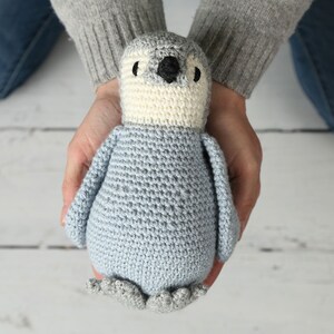 May include: A crocheted penguin toy with a light blue body, white belly, and gray feet. The penguin is being held in the hands of a person.