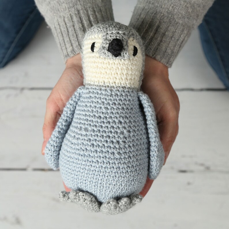 May include: A crocheted penguin toy with a light blue body, white belly, and gray feet. The penguin is being held in the hands of a person.