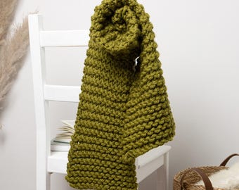 May include: A chunky knit scarf in a muted olive green color is draped over the back of a white wooden chair. The scarf's thick texture and warm hue suggest cozy comfort. A stack of books and a woven basket are also visible.