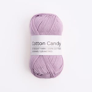 May include: A skein of light purple DK weight yarn made of 100% cotton. A white paper label wraps around the center, printed with the text Cotton Candy, DK WEIGHT YARN, 100% COTTON, 50GRAMS, and 125 METRES in a clear, black sans-serif font.