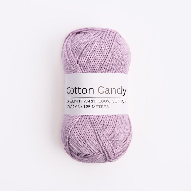 May include: A skein of light purple DK weight yarn made of 100% cotton. A white paper label wraps around the center, printed with the text Cotton Candy, DK WEIGHT YARN, 100% COTTON, 50GRAMS, and 125 METRES in a clear, black sans-serif font.