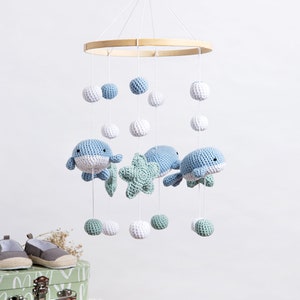 May include: A hanging mobile featuring crocheted light blue and white whales, a mint green starfish, and small crocheted spheres in shades of blue, white, and mint green suspended from a circular wooden hoop by thin white strings.