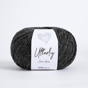 K&ouml;nnte beinhalten: Ein Kn&auml;uel dunkel anthrazitfarbenes Aran-Garn mit einem wei&szlig;en Papieretikett. Das Etikett zeigt ein Herzsymbol, den Text Wool London, Utterly, Aran Yarn sowie 50g approx. 100m approx. Das Garn hat eine texturierte, leicht gesprenkelte Optik.