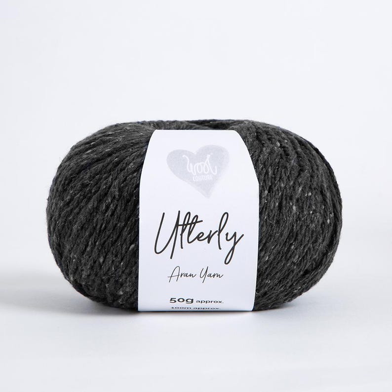 K&ouml;nnte beinhalten: Ein Kn&auml;uel dunkel anthrazitfarbenes Aran-Garn mit einem wei&szlig;en Papieretikett. Das Etikett zeigt ein Herzsymbol, den Text Wool London, Utterly, Aran Yarn sowie 50g approx. 100m approx. Das Garn hat eine texturierte, leicht gesprenkelte Optik.