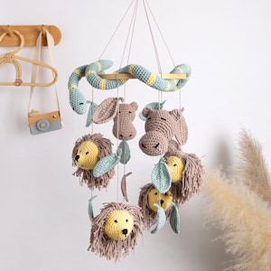 May include: A hanging mobile featuring crocheted figures of lions with yellow faces and brown manes, brown hippos, and a long light blue and yellow striped snake. The figures are suspended from a wooden crossbar by thin strings.