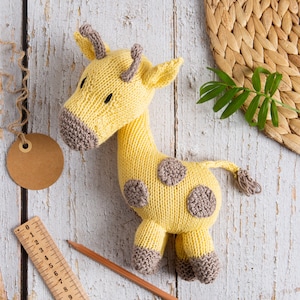 May include: A yellow knitted giraffe toy with brown spots, a brown muzzle, and two small brown horns. The giraffe is positioned next to a wooden ruler showing measurements in centimetres, a brown pencil, and a blank circular brown paper tag.