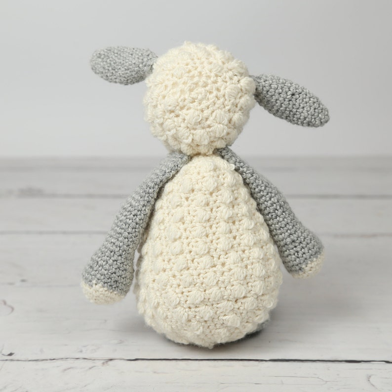 May include: A crocheted stuffed lamb toy with a white body and gray ears. The lamb is standing on a white wooden surface.