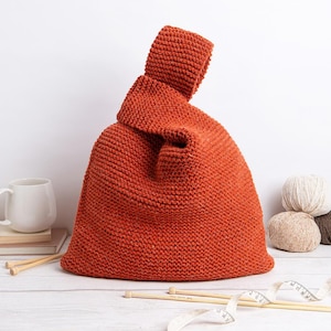 Puede incluir: Un bolso tejido a mano de color óxido, con un asa anudada única. El bolso está hecho de hilo grueso y tiene un aspecto texturizado. También se ven agujas de tejer, ovillos de lana y una cinta métrica.