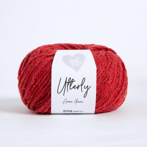 K&ouml;nnte beinhalten: Ein Kn&auml;uel rotes Aran-Garn mit einem wei&szlig;en Papieretikett, das den Text Wool Couture, Utterly, Aran Yarn und 50g approx. tr&auml;gt. Das Garn hat eine strukturierte, leicht melierte Optik mit subtilen Farbvariationen im Rot der Fasern.