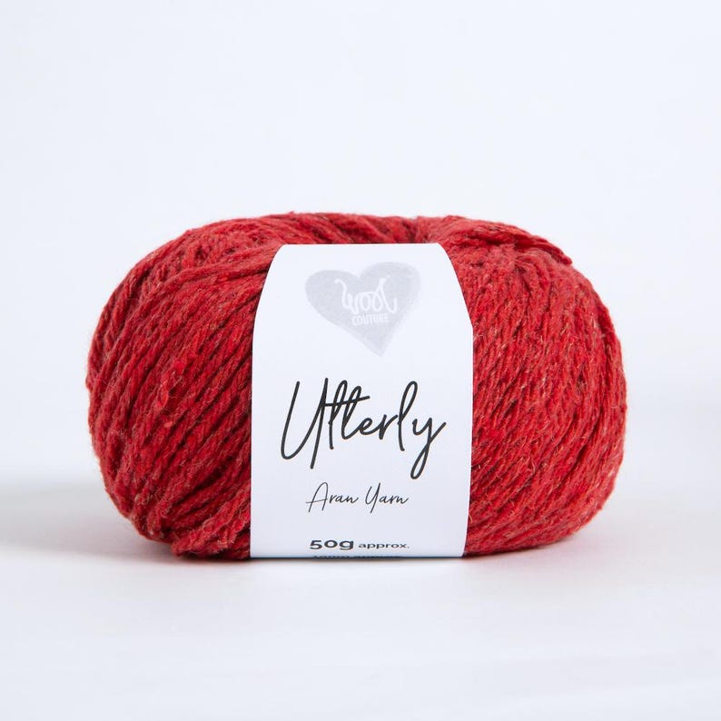 K&ouml;nnte beinhalten: Ein Kn&auml;uel rotes Aran-Garn mit einem wei&szlig;en Papieretikett, das den Text Wool Couture, Utterly, Aran Yarn und 50g approx. tr&auml;gt. Das Garn hat eine strukturierte, leicht melierte Optik mit subtilen Farbvariationen im Rot der Fasern.