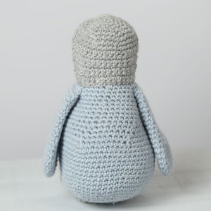 May include: A crocheted penguin toy with gray and light blue yarn. The penguin is standing upright with its wings out to the sides.