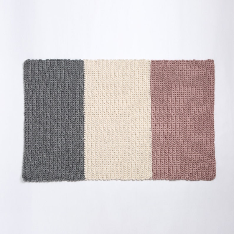 May include: A rectangular crocheted rug with three vertical stripes in gray, white, and pink.