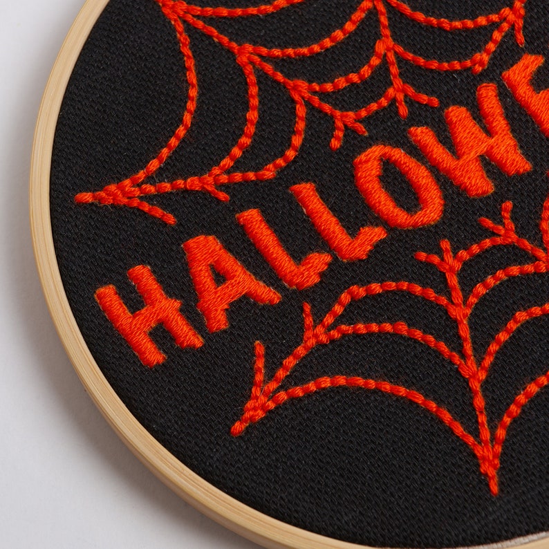 May include: Embroidered Halloween hoop art with a black background and orange thread. The word "HALLOWEEN" is stitched in a bold font with a spiderweb design behind it.
