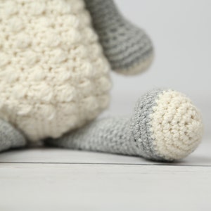 May include: A crocheted stuffed animal toy in the shape of a sheep. The sheep is white with gray accents on the legs and head. The sheep is sitting on a white surface.