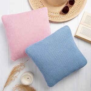 Könnte beinhalten: Zwei quadratische Strickkissen, eines in Hellrosa und eines in Hellblau, liegen auf einer weißen Oberfläche. Ein Sonnenhut aus Stroh, eine Sonnenbrille und ein offenes Buch sind ebenfalls vorhanden, was auf ein entspannendes Thema hindeutet. Eine brennende Kerze und getrocknete Pflanzen vervollständigen die Szene.