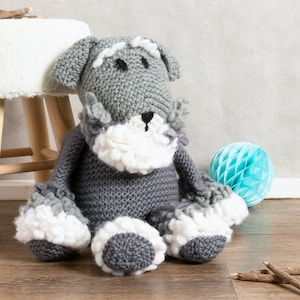 May include: A knitted dog plush toy in grey and white, featuring floppy ears, a white beard, and black eyes. The toy is positioned on a wooden floor. A white stool and a blue honeycomb ball are visible in the background.