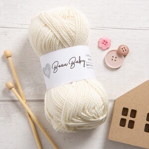 May include: A ball of white yarn with a label that reads "Beau Baby" and a heart logo. The yarn is next to two wooden knitting needles, a pink button, and a brown button.