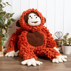 Pode incluir: Um brinquedo de pelúcia de orangotango de malha laranja e marrom com mãos e pés brancos. O brinquedo está sentado em um chão de madeira.