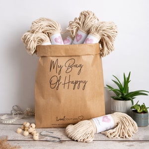 May include: A brown paper bag filled with natural cotton rope, labeled "My Bag Of Happy" and "woolCOUTURE". The bag is sitting on a wooden surface with additional spools of rope, wooden beads, and measuring tape.