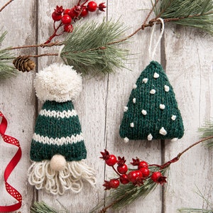 May include: A knitted gnome ornament with a dark green and white striped hat topped with a white pom-pom and a white yarn beard, alongside a triangular dark green knitted tree ornament featuring small white tufts and a white hanging loop.