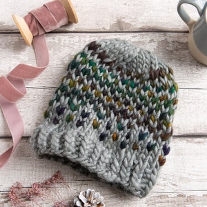 Könnte beinhalten: Eine graue Strickmütze mit einem bunten Muster aus grünem, braunem und violettem Garn. Die Mütze ist aus einem dicken Garn gefertigt und hat eine dicke, warme Textur.