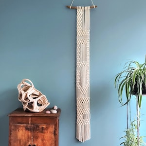 May include: A white macrame wall hanging with a geometric pattern. The hanging is made of cotton cord and is attached to a wooden dowel. The hanging is against a light blue wall.