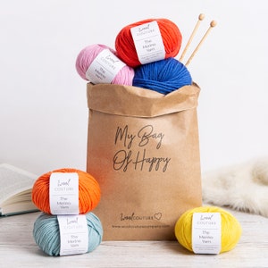 May include: A brown paper bag with the text "My Bag Of Happy" printed on it. The bag is filled with six balls of yarn in various colours, including red, pink, blue, orange, yellow, and green. The yarn is labelled "Wool Couture The Merino Yarn".