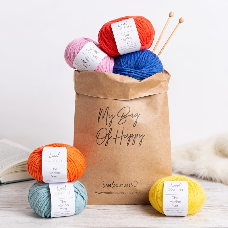 May include: A brown paper bag with the text "My Bag Of Happy" printed on it. The bag is filled with six balls of yarn in various colours, including red, pink, blue, orange, yellow, and green. The yarn is labelled "Wool Couture The Merino Yarn".