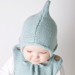 Puede incluir: Un bebé con un gorro de punto azul claro con capucha y punta puntiaguda. El gorro está hecho de un hilo suave y texturizado y tiene un diseño sencillo.