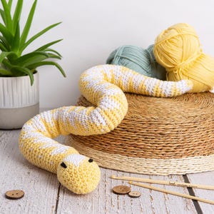 Pode incluir: Um brinquedo de cobra em crochê com um padrão repetitivo de fio amarelo e branco. A cobra tem dois pequenos olhos pretos e está posicionada perto de duas agulhas de crochê de madeira, dois botões castanhos e dois novelos de lã.