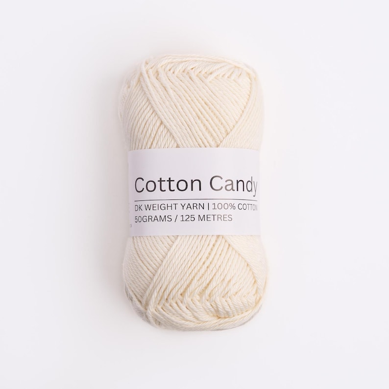 May include: A skein of off-white DK weight yarn made of 100% cotton. The label reads Cotton Candy, DK WEIGHT YARN, 100% COTTON, 50GRAMS / 125 METRES. The yarn is wound into a compact, oval-shaped ball with visible twisted strands of fiber.