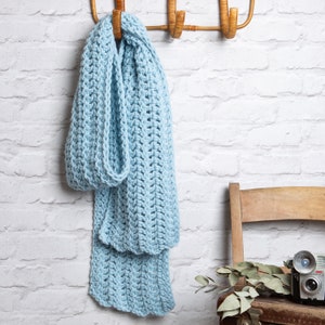 Puede incluir: Una bufanda de crochet azul claro colgada de un gancho de madera contra una pared de ladrillo blanco.
