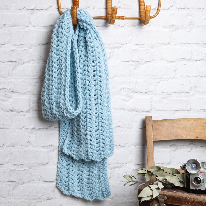 Puede incluir: Una bufanda de crochet azul claro colgada de un gancho de madera contra una pared de ladrillo blanco.