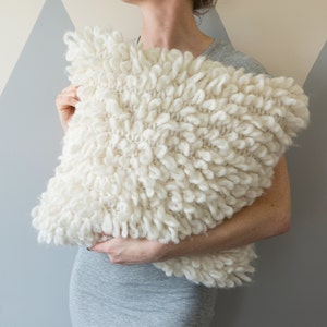 May include: A white knitted throw pillow with a fluffy, textured surface. The pillow is held by a person's hands.