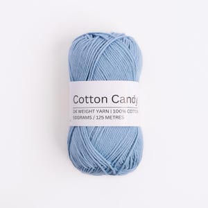 May include: A skein of light blue DK weight yarn wrapped in a white paper label that reads Cotton Candy, DK WEIGHT YARN, 100% COTTON, 50 GRAMS, 125 METRES. The yarn has a smooth, twisted texture and is presented in a compact, oval-shaped bundle.